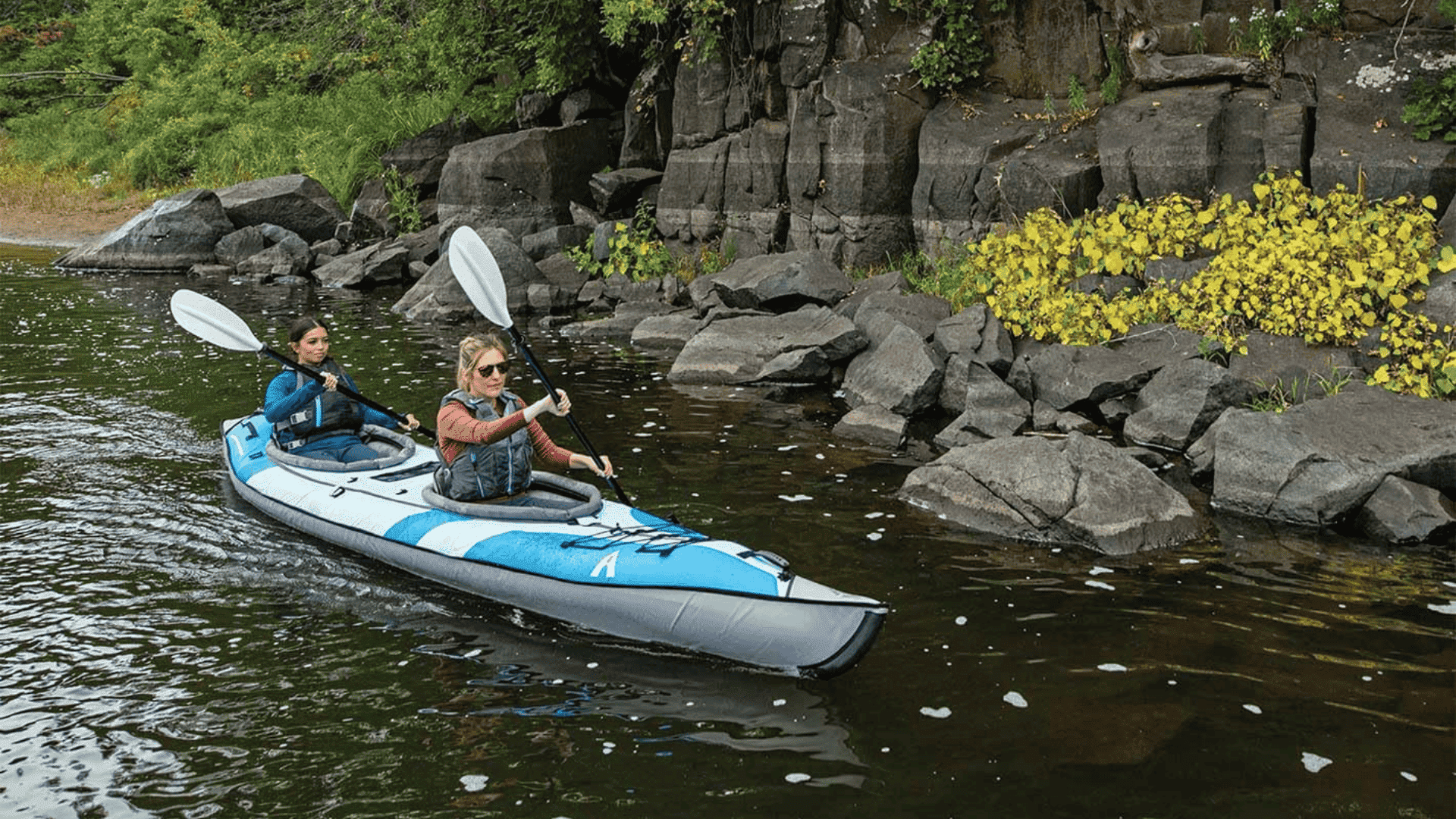 Two people paddling a blue and grey Advanced Elements AdvancedFrame Convertible tandem kayak near a rocky shoreline.