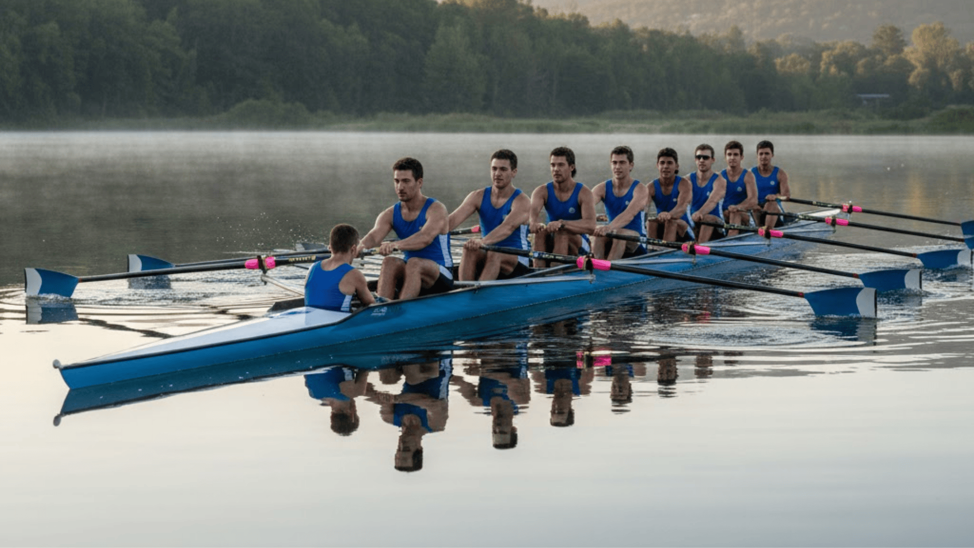 A rowing team of eight athletes in blue uniforms sits in a long racing shell on calm water at dawn