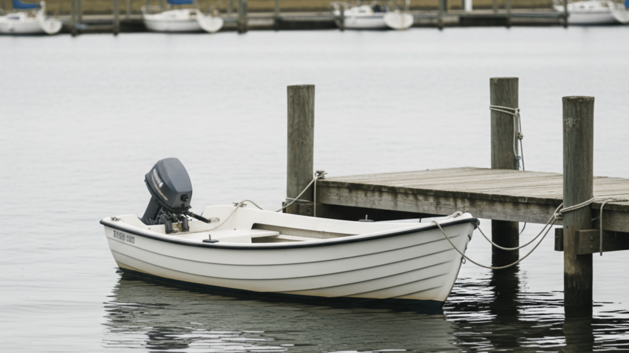 A small white motorboat with an outboard engine is tied to a wooden pier on a calm body of water