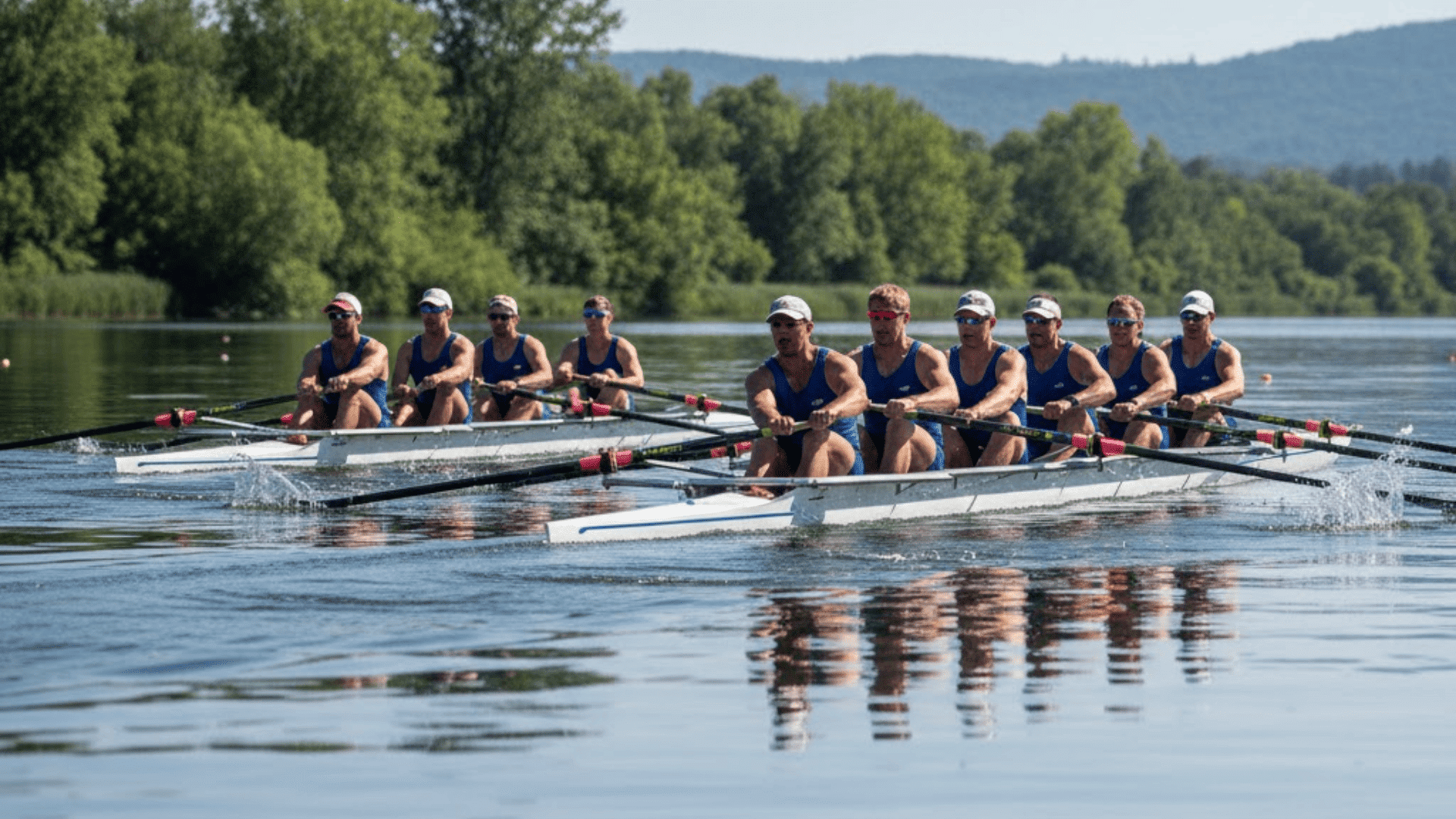 Two rowing crews race side by side on a river, with eight rowers in blue uniforms paddling in sync