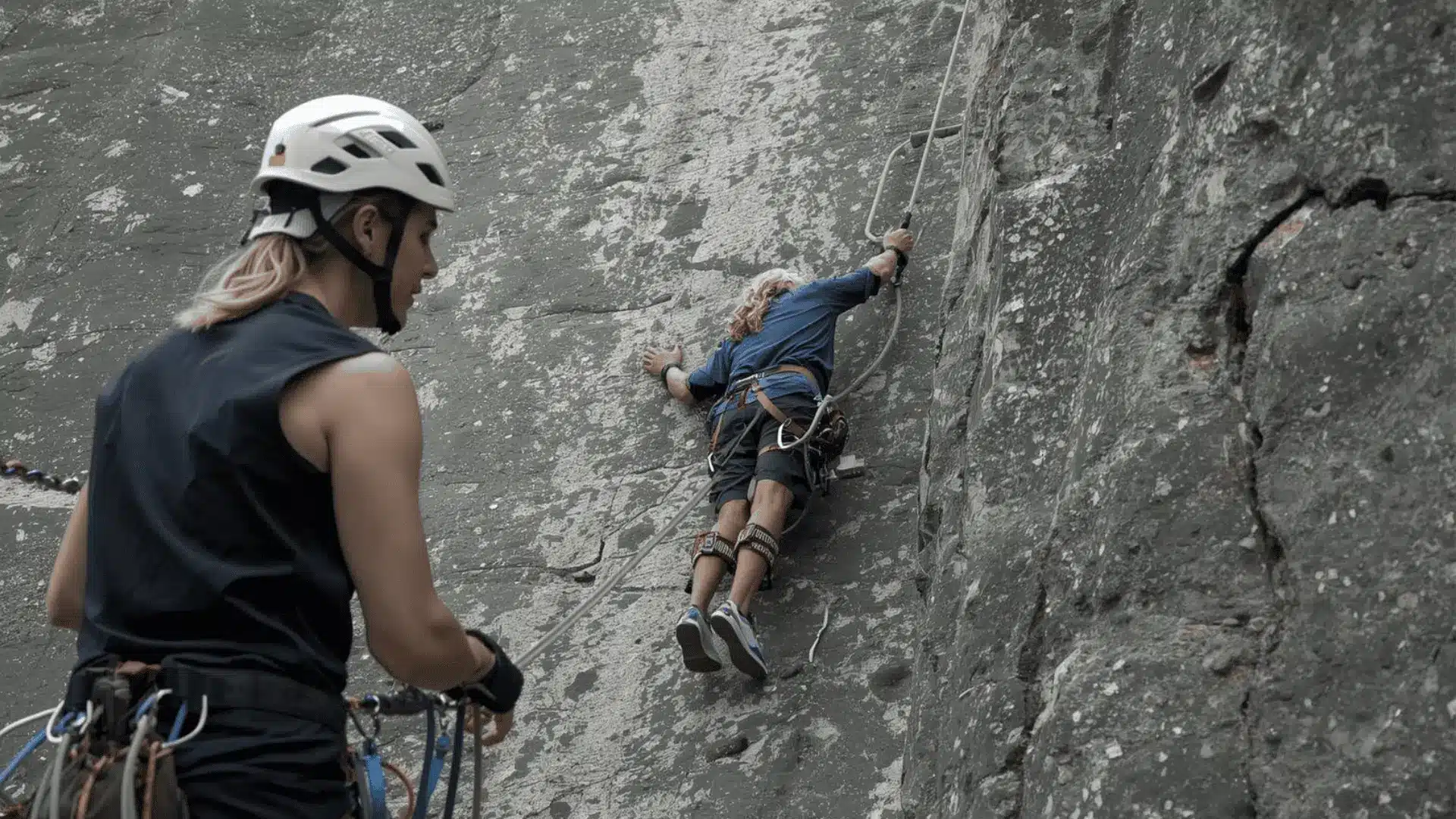 A climber ascends a rock wall with a guide nearby, illustrating the Via Ferrata climbing adventure with fixed holds and cables