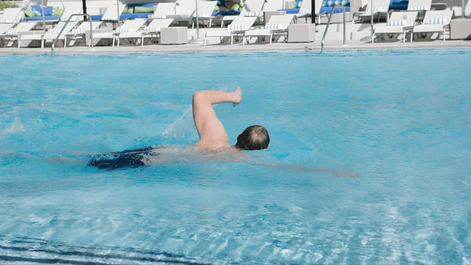 Man swimming freestyle in an outdoor pool with lounge chairs and umbrellas visible on the deck behind him