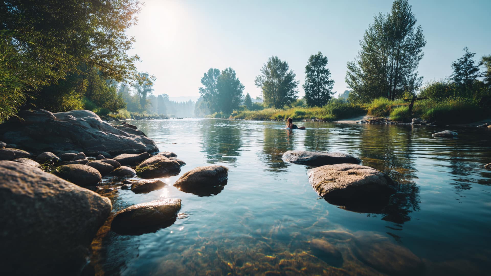 Clear river with smooth rocks in the foreground, sunlight reflecting on calm running water, surrounded by green trees under a bright blue sky.