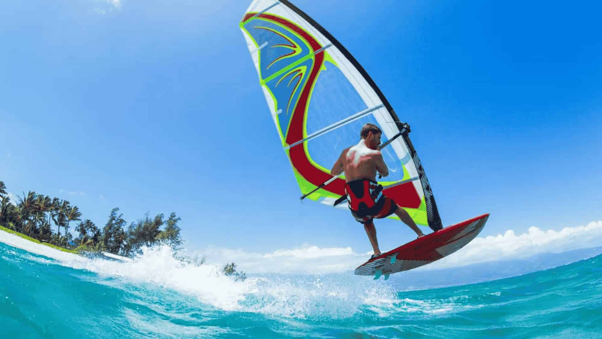 Windsurfer performing a jump over a wave on turquoise water near a tropical shoreline under a clear blue sky.