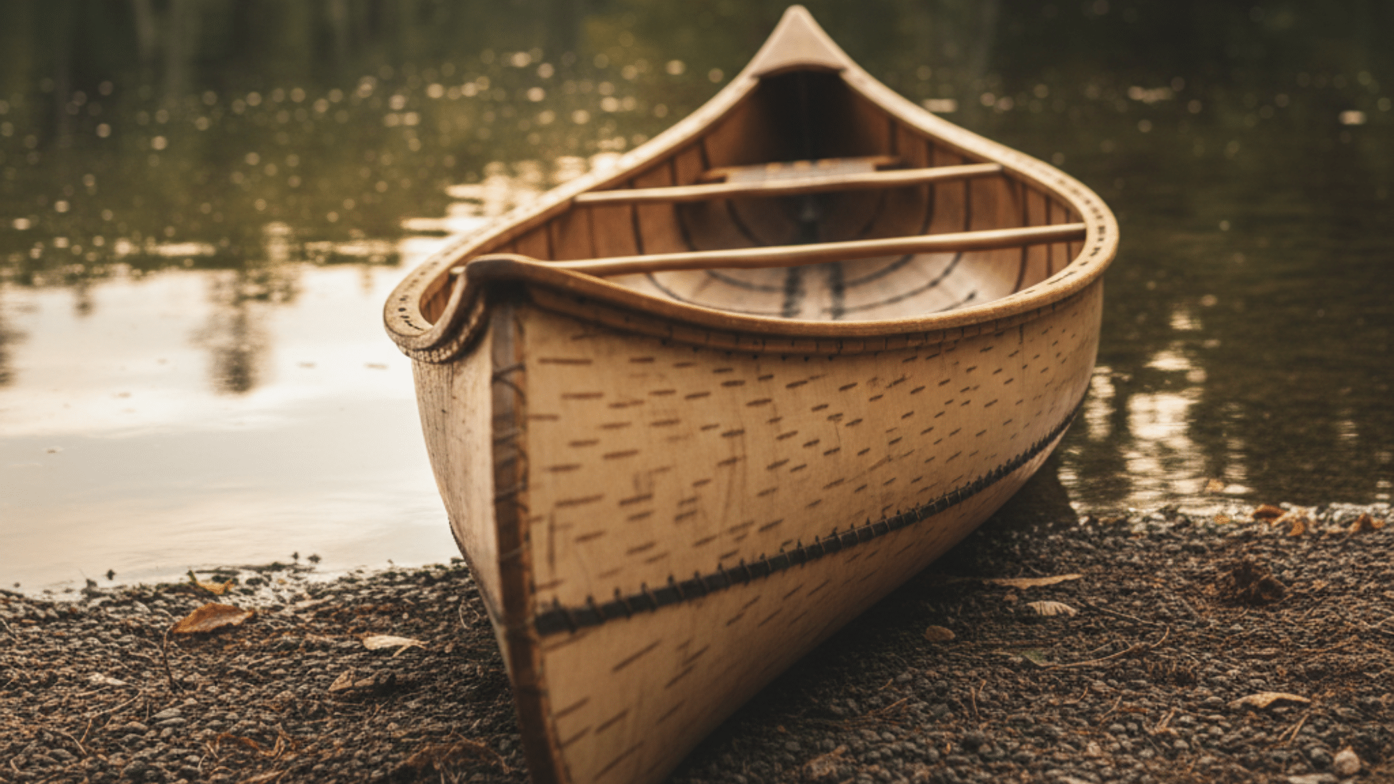 Traditional wooden birchbark canoe resting on a lakeshore beside calm water with forest reflections.