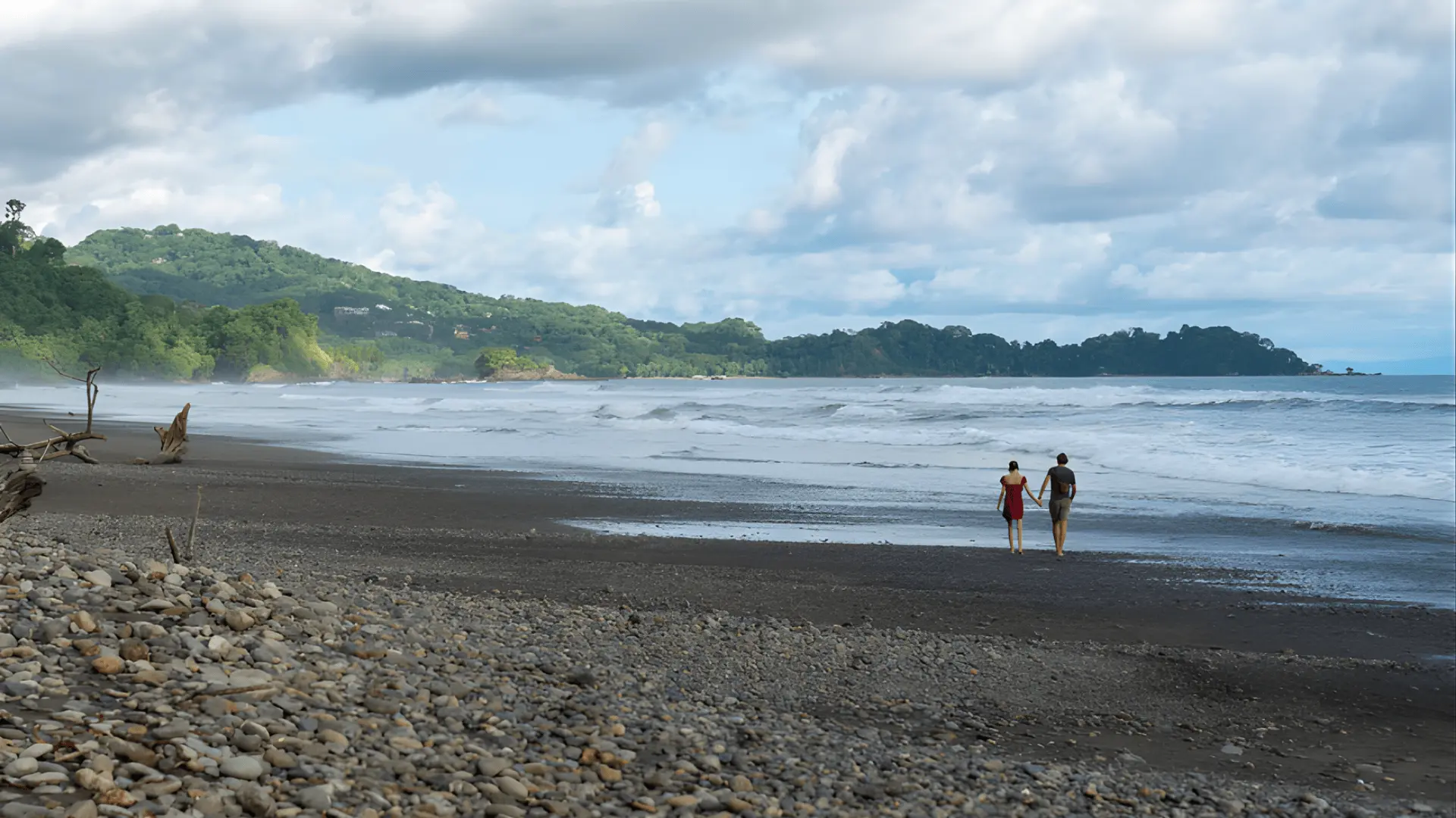 Two people walking along a rocky beach in Dominical, surrounded by lush jungle and the Pacific Ocean waves.