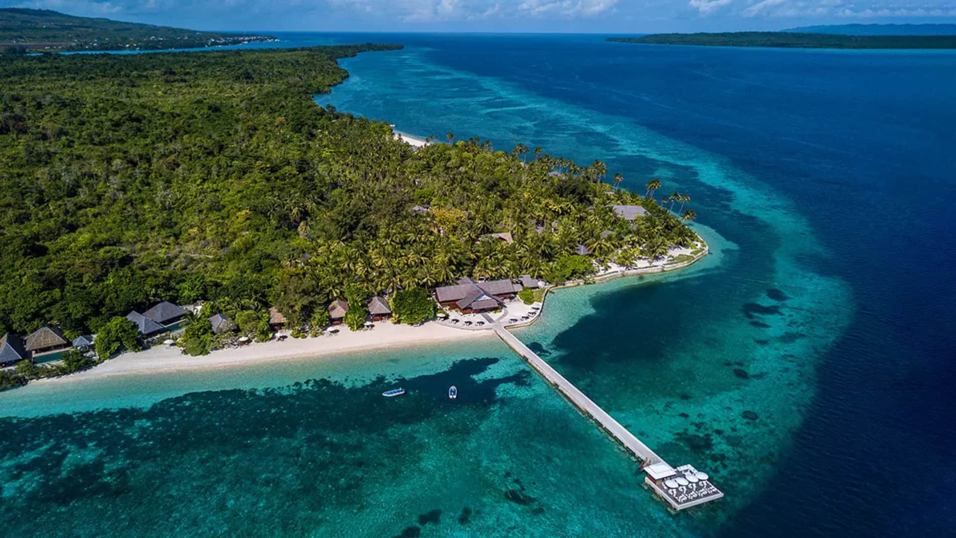 Aerial view of a tropical island resort with a long pier over clear turquoise water.
