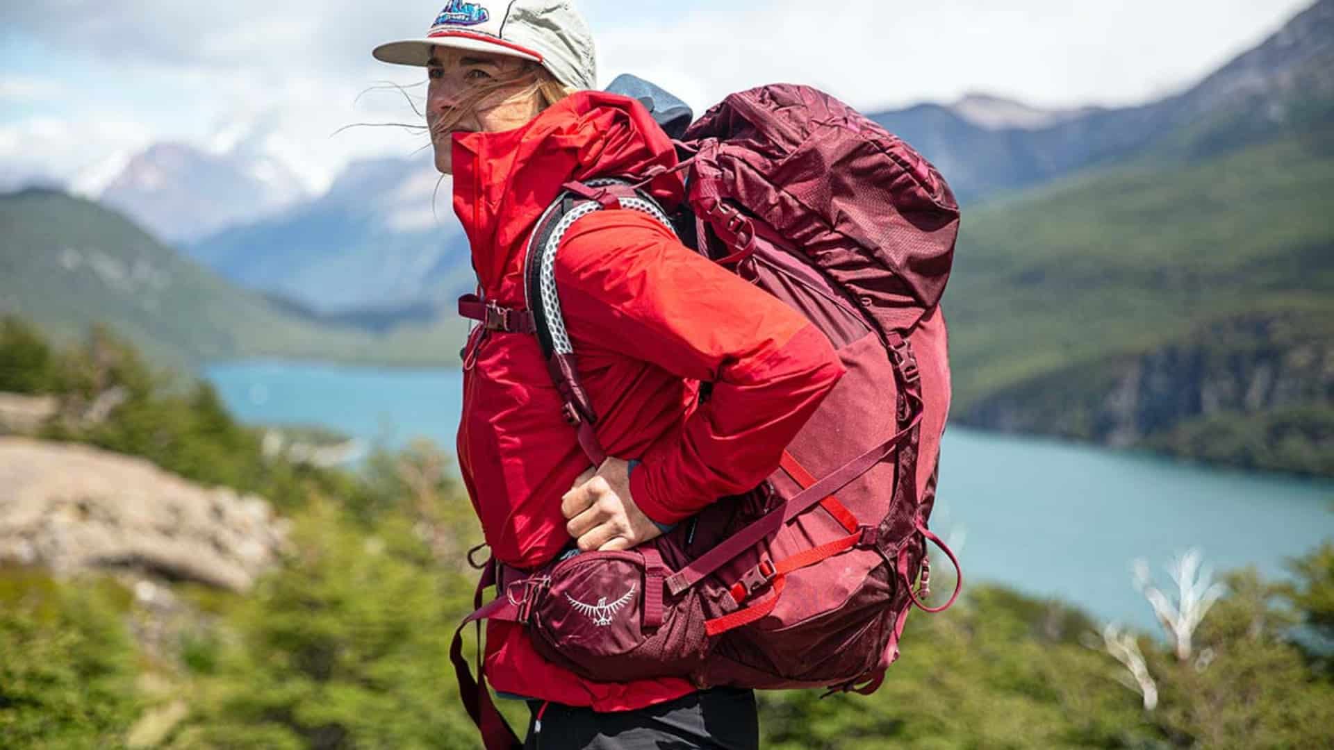 Woman hiking on mountain trail wearing Osprey Aura AG 65 backpack with lake and mountain landscape in background.