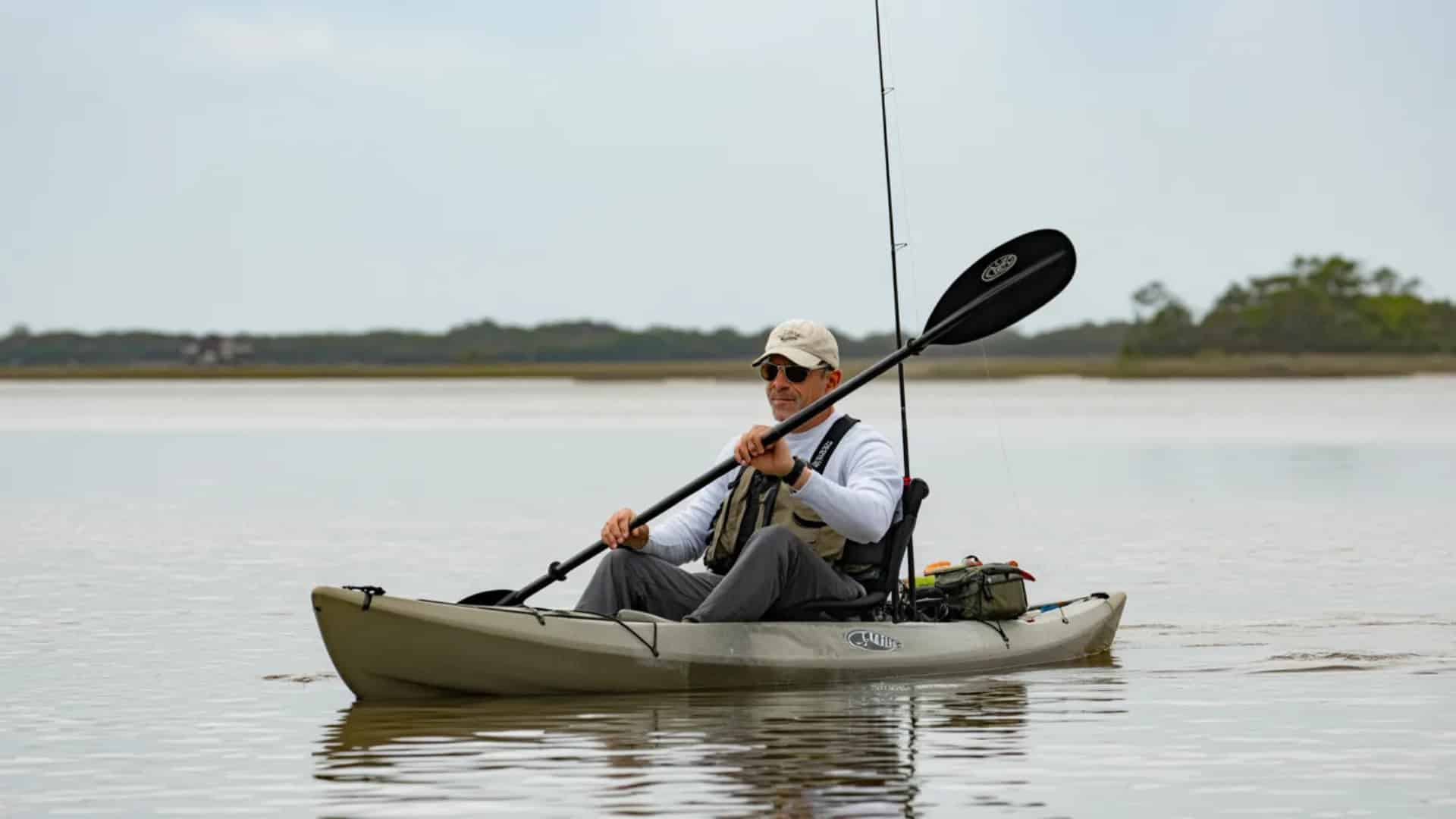 Angler paddling a fishing kayak across calm water under a soft gray morning sky