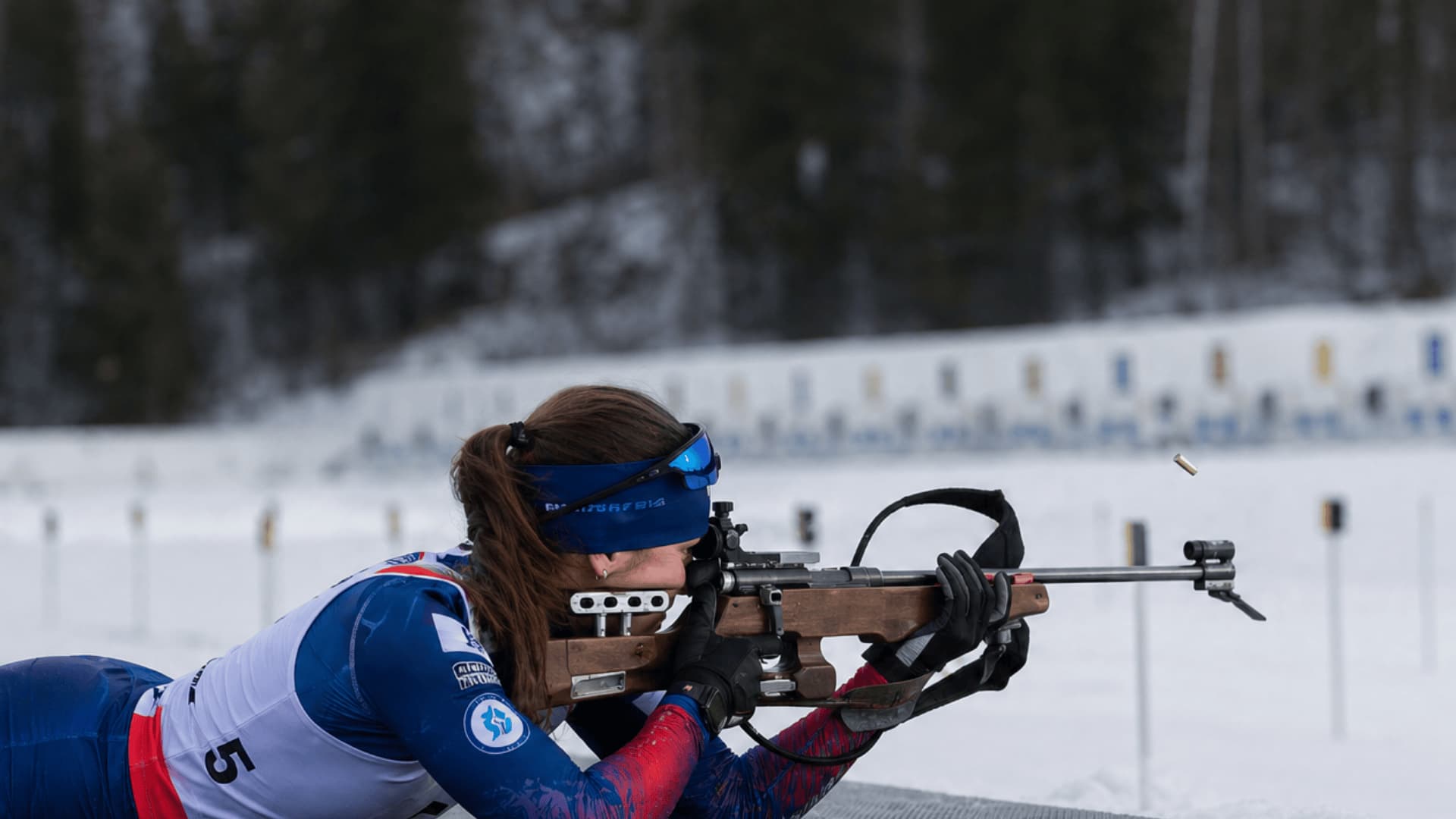 Biathlon athlete lying prone on a snowy shooting range, aiming a rifle at a distant target in a winter forest setting