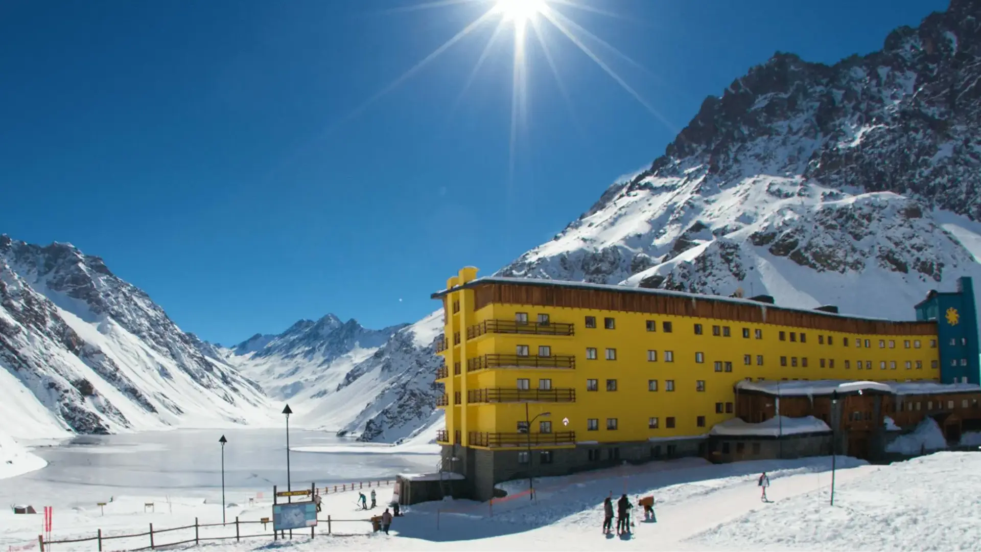 Bright yellow alpine hotel beside a frozen mountain lake with snowy peaks and skiers under a sunny blue sky