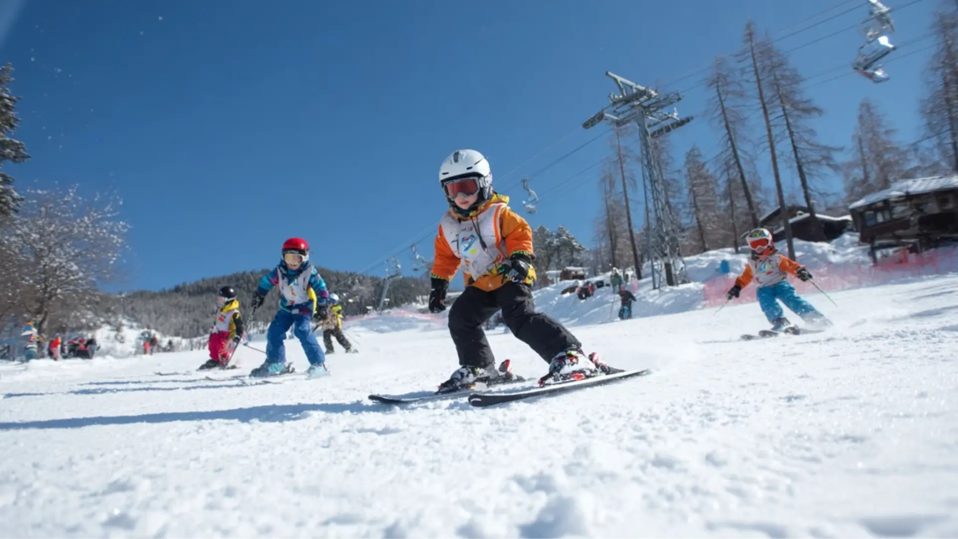 Children skiing on a groomed snowy slope at a ski resort with ski lifts and trees under a clear blue sky