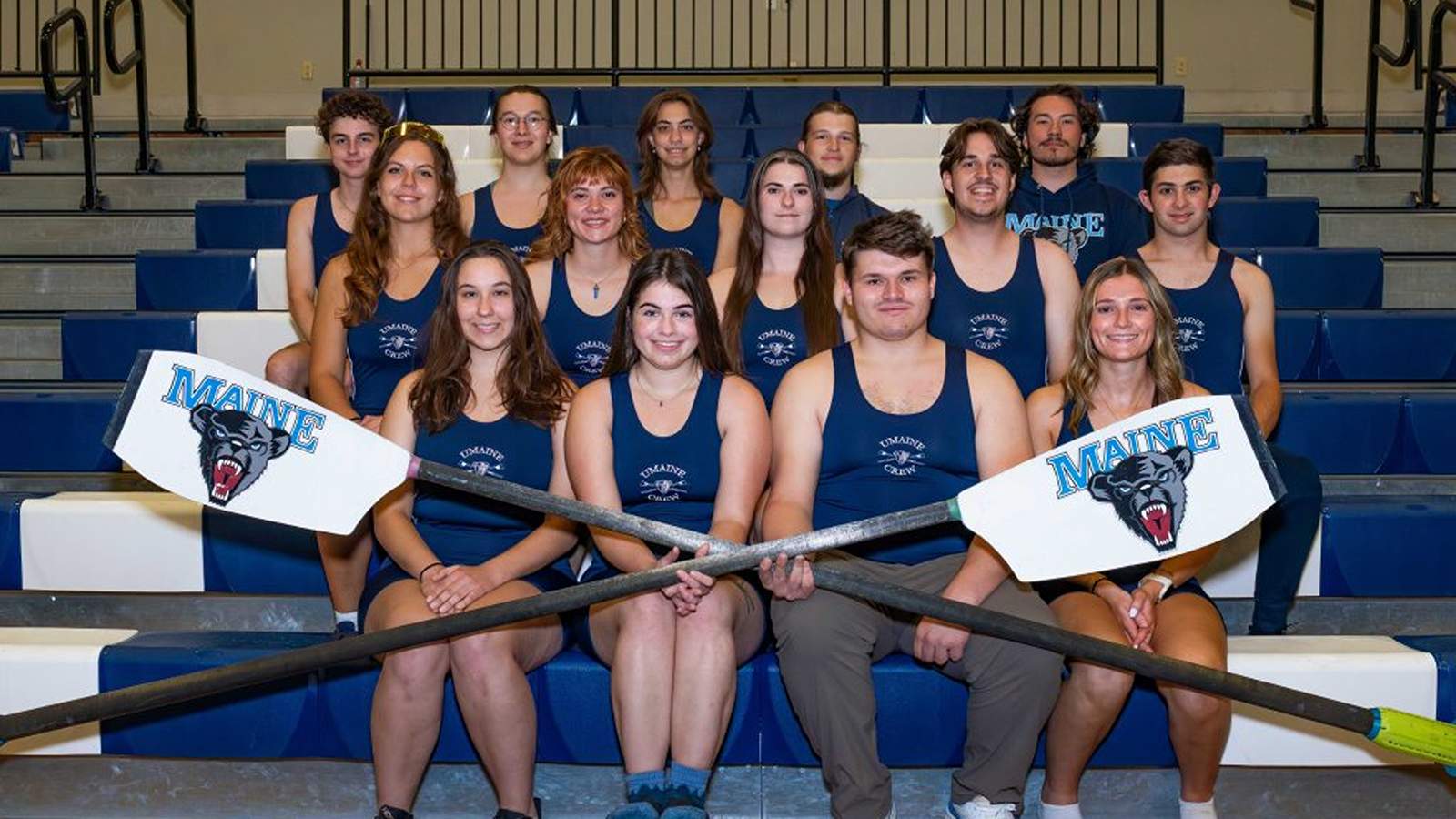 College rowing team posing indoors with oars, wearing team uniforms on bleachers for a group photo_