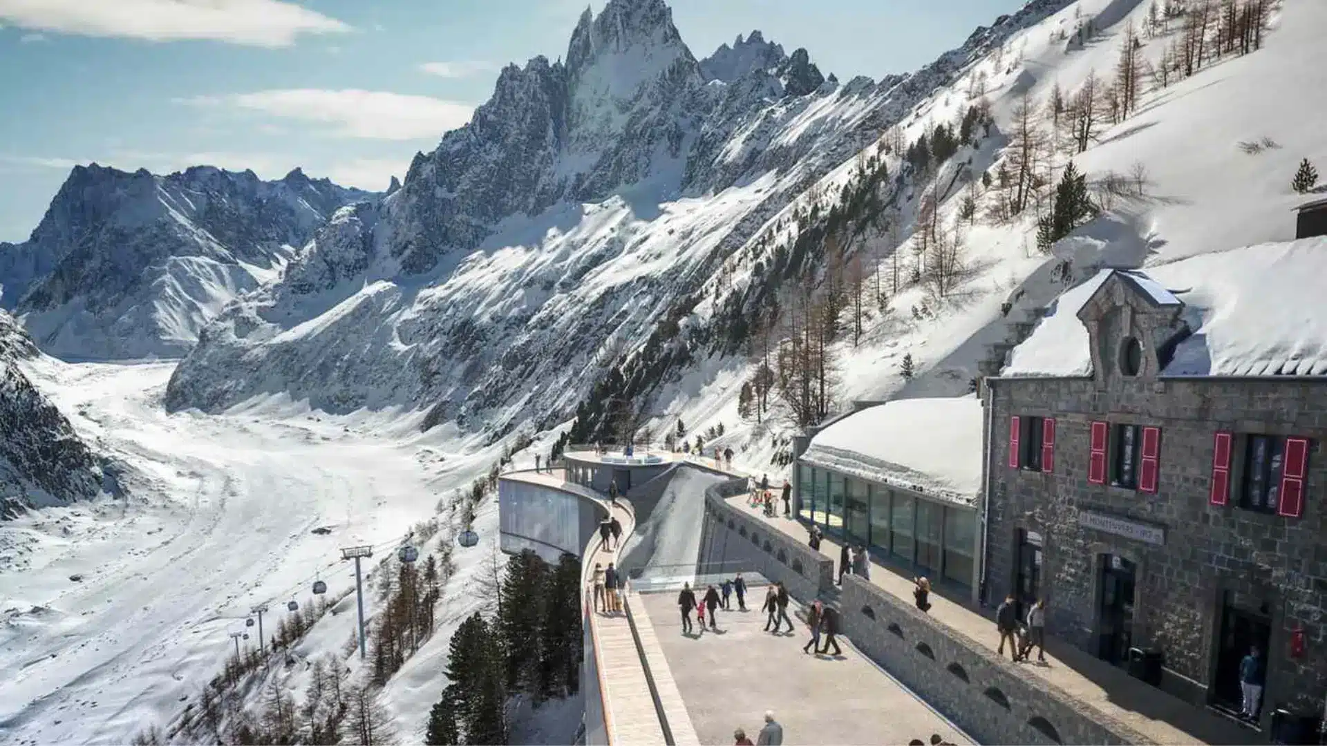 Mountain viewpoint terrace at a snowy alpine resort with visitors walking near a stone building, overlooking dramatic jagged peaks and a glacier valley
