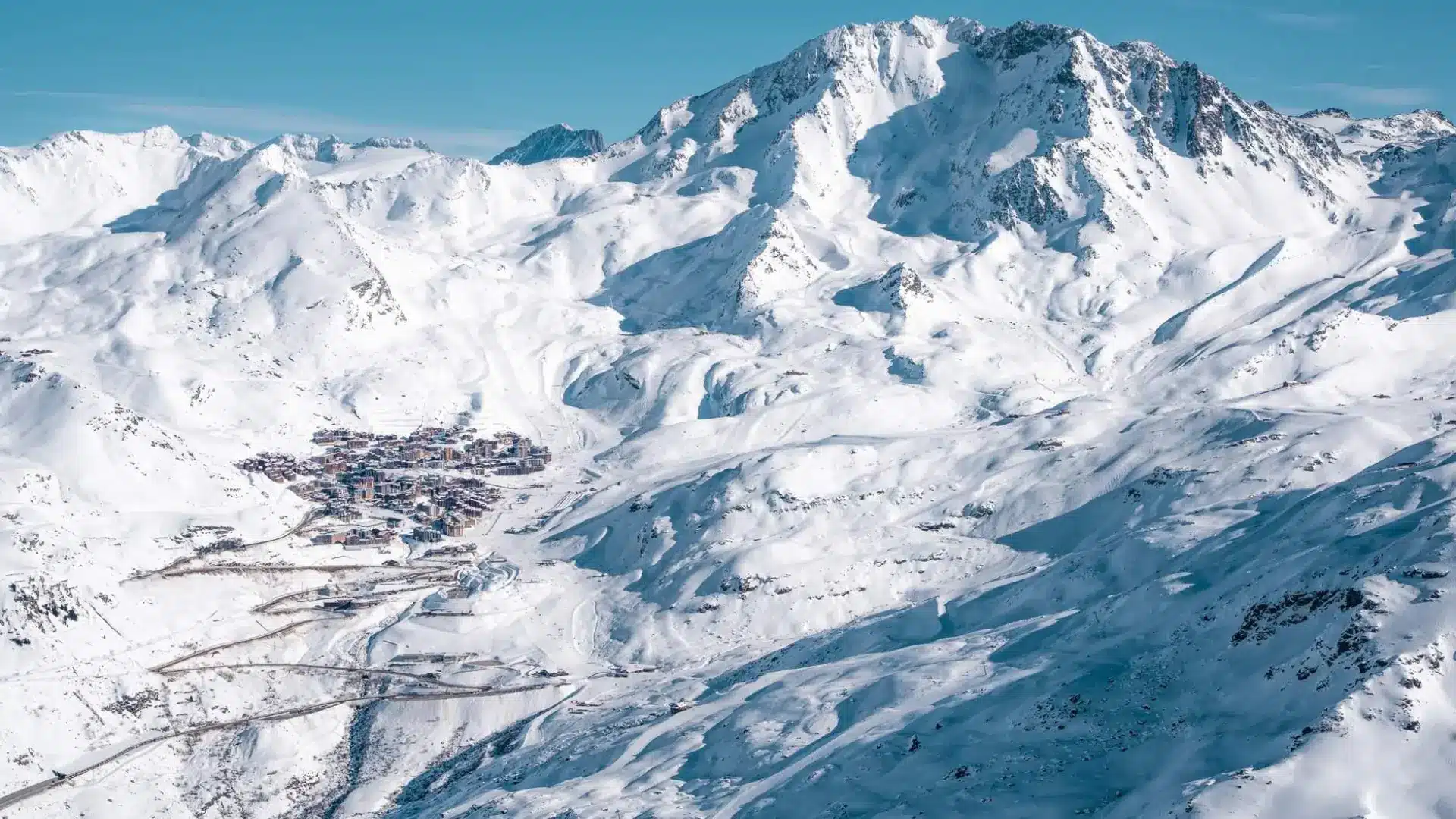 Panoramic view of a snowy alpine ski resort village nestled in a mountain valley, surrounded by vast white slopes and towering peaks under a clear blue sky