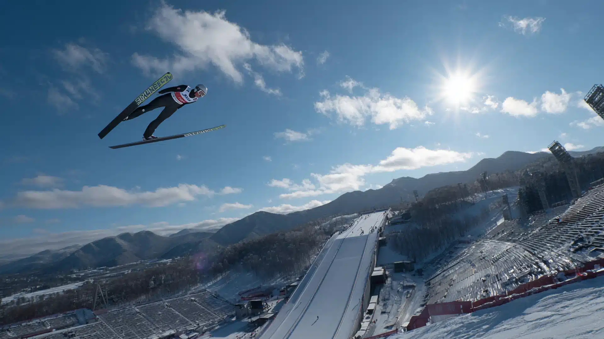 Ski jumper soaring above long ramp with snowy stadium and mountains behind now