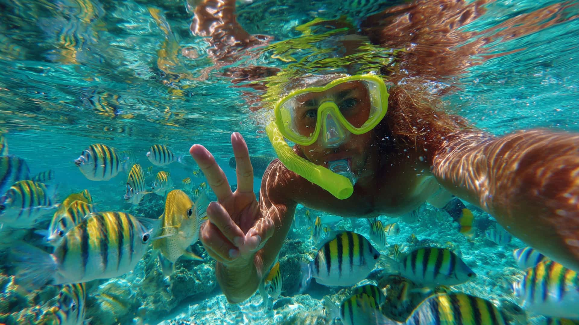 Snorkeler takes a selfie underwater with tropical fish and flashes a peace sign
