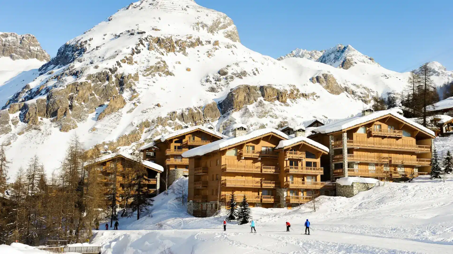 Snow-covered alpine chalets with skiers on slopes and rugged mountain peaks under a clear blue sky