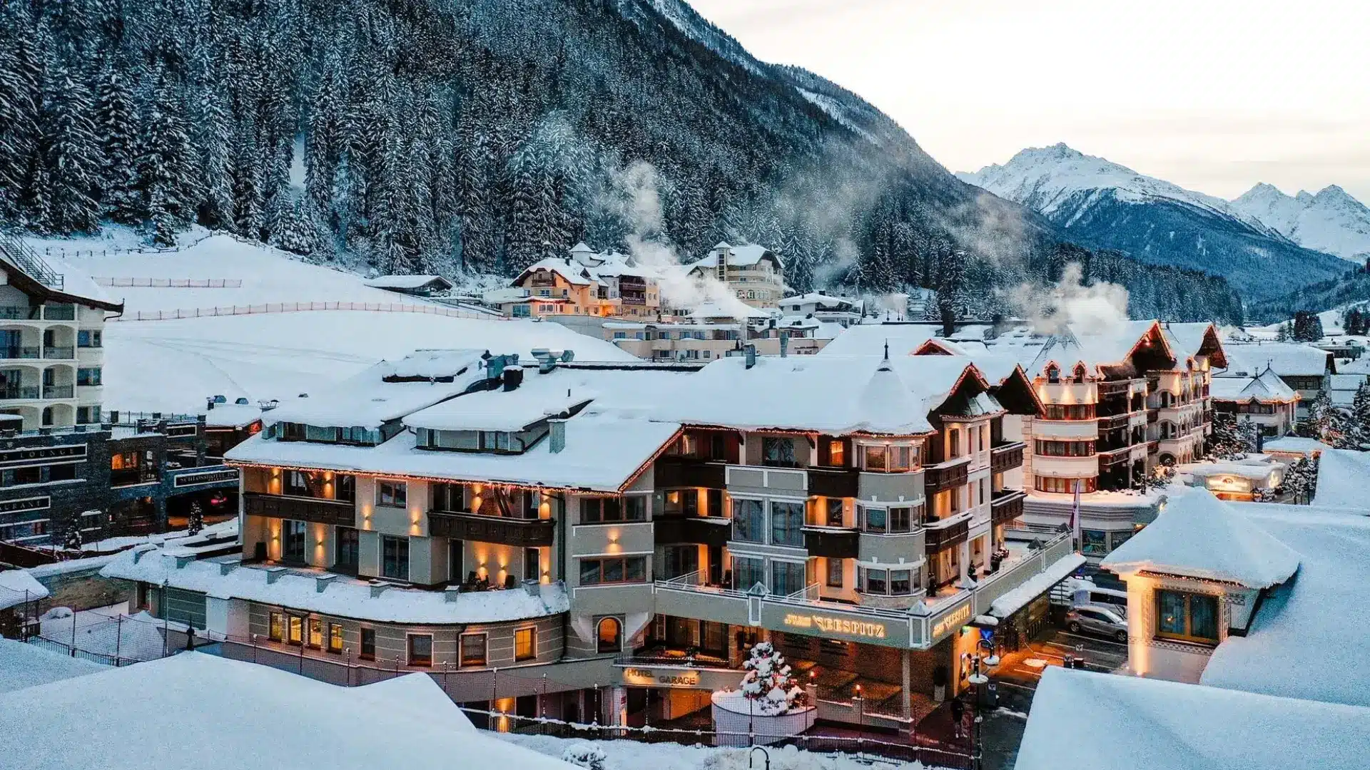 Snow-covered alpine resort village with cozy hotels, glowing lights, and forested mountains in the background