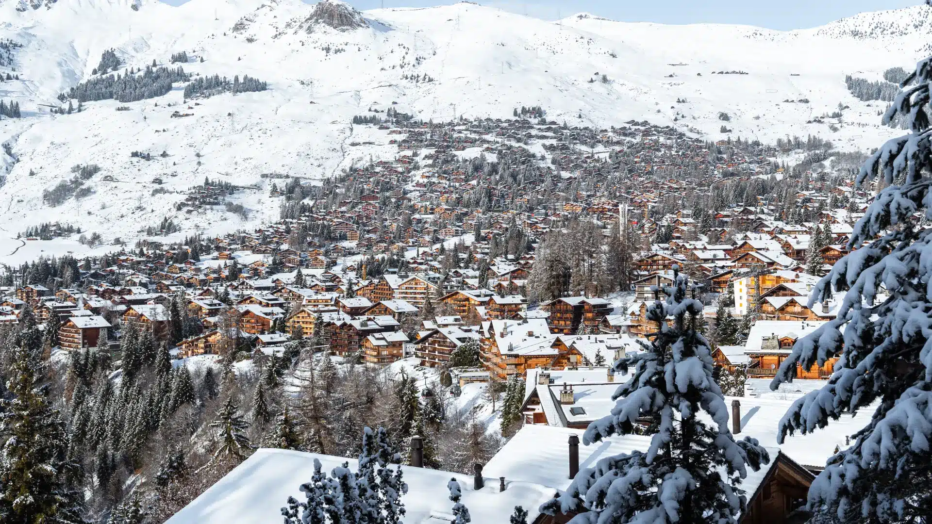 Snow-covered alpine ski resort town with wooden chalets, forested slopes, and wide mountain terrain under clear skies