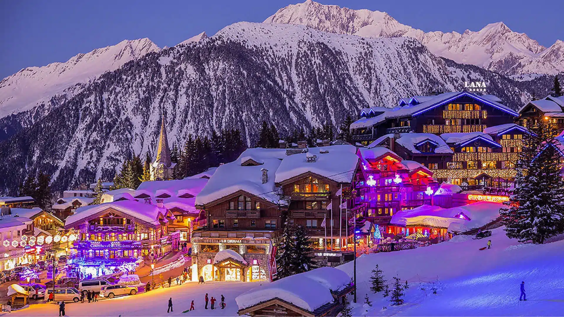 Snow-covered alpine ski resort village at night with glowing lights, chalets, and mountain peaks in the background