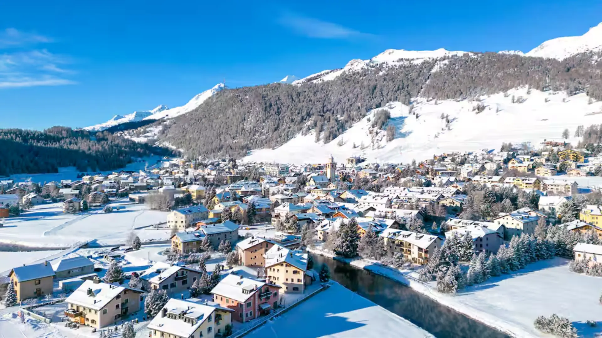 Snow-covered alpine town with colorful houses, river, and surrounding mountains under a clear blue sky