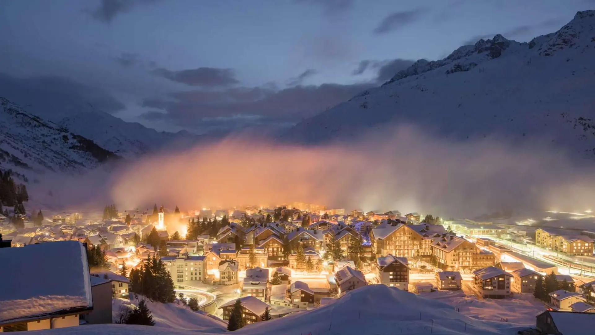 Snow-covered alpine village at dusk with glowing lights, mist over the town, and surrounding mountain slopes