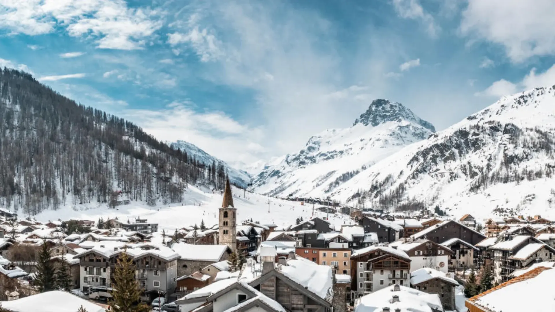 Snow-covered alpine village in the French Alps with chalet-style houses, church tower, and surrounding mountain peaks