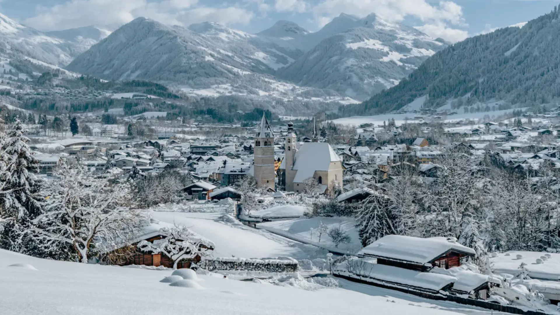 Snow-covered alpine village with church towers, chalets, and surrounding mountains in a peaceful winter landscape