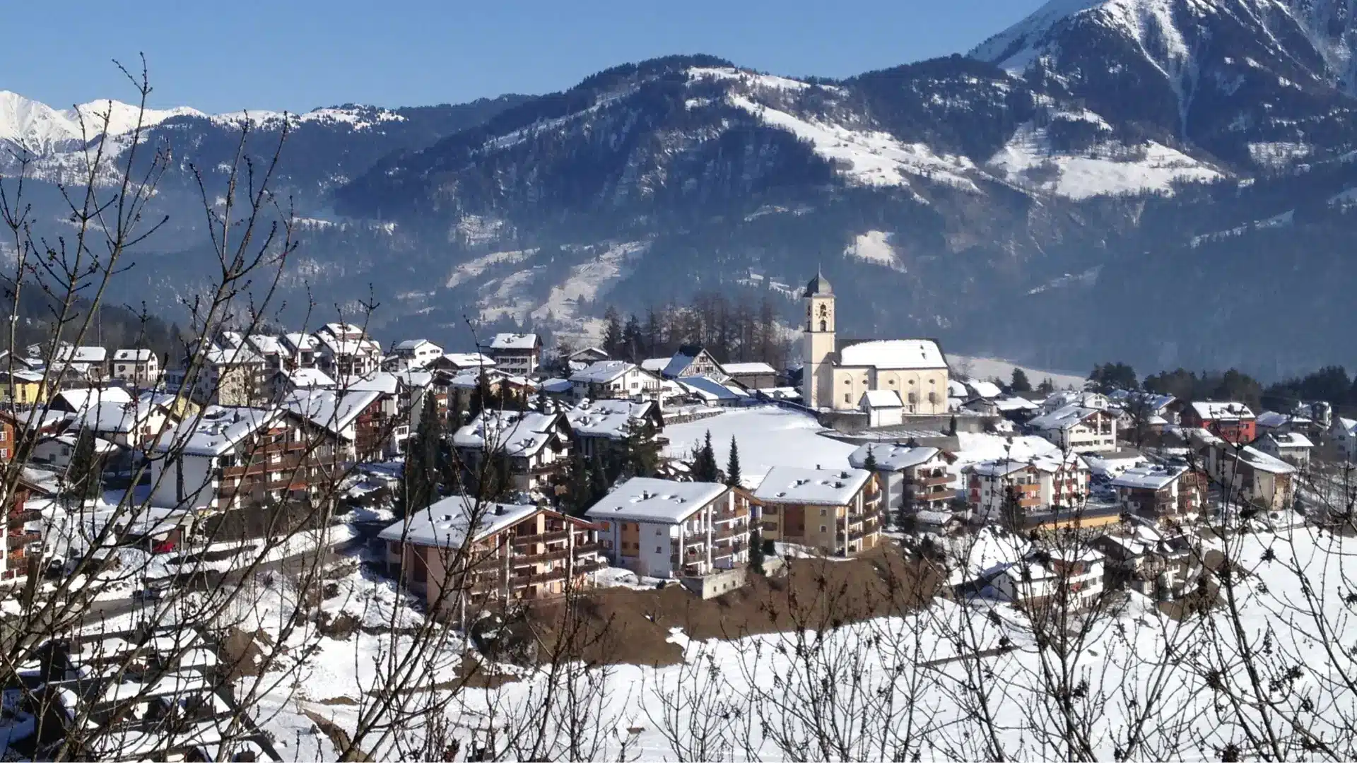 Snow-covered alpine village with clustered houses and a church at its center, set against forested mountains under a clear blue sky