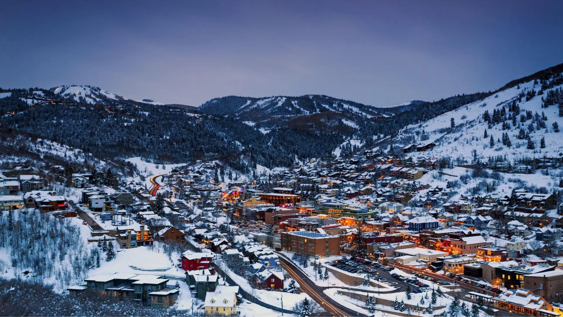 Snow-covered mountain town at dusk with glowing streets, ski slopes, and forested hills in the background
