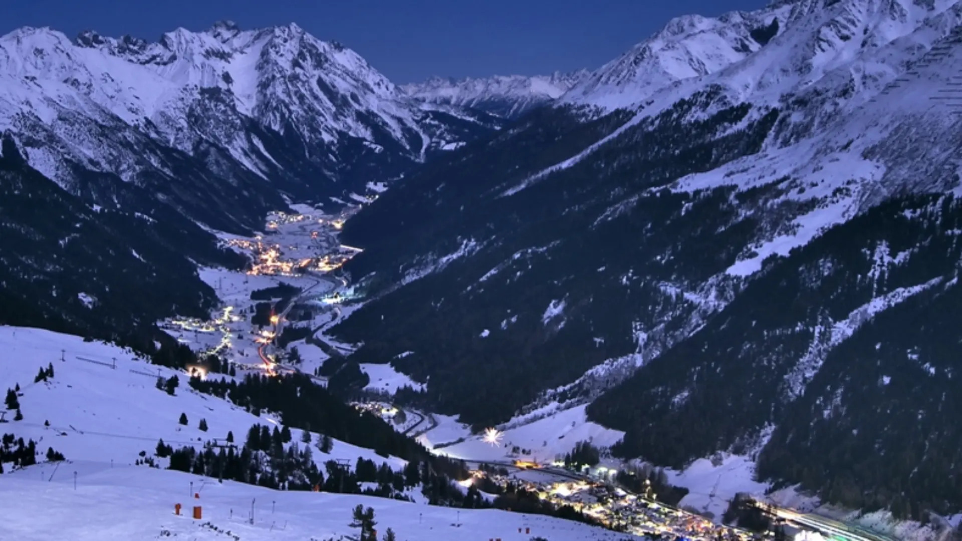 Snow-covered mountain valley at dusk with a lit alpine village, winding roads, and surrounding high peaks