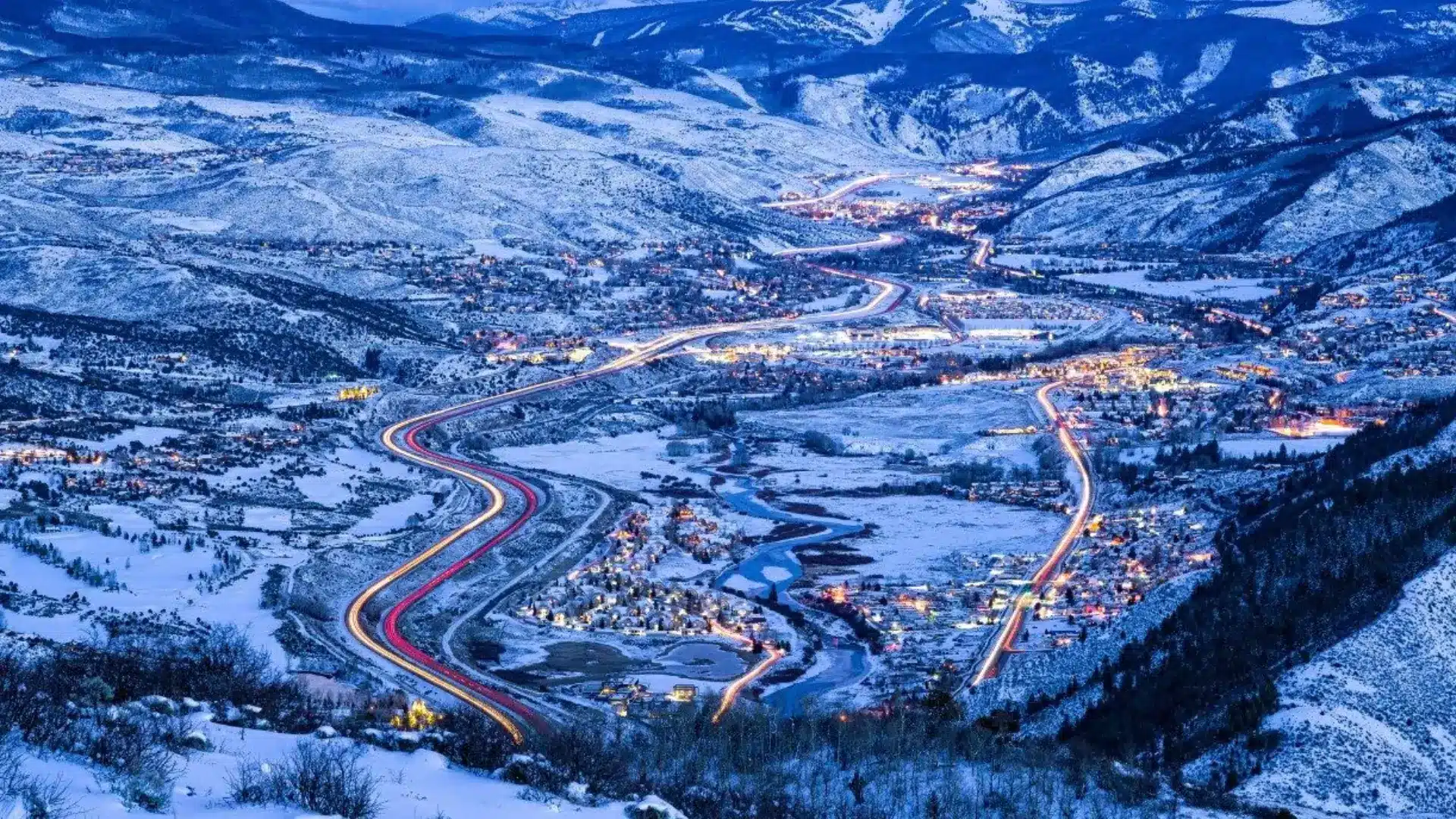 Snow-covered mountain valley with winding highway lights, small towns, and rolling hills under a winter sky