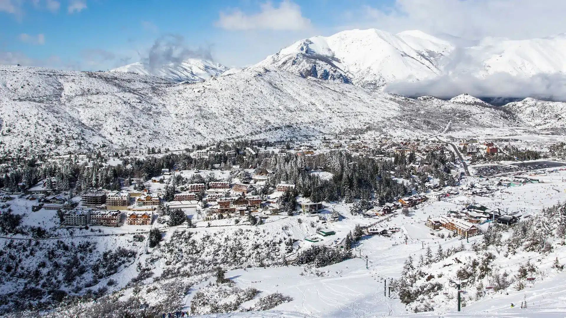 Snow-covered mountain village with ski slopes, pine trees, and scattered buildings beneath cloudy peaks