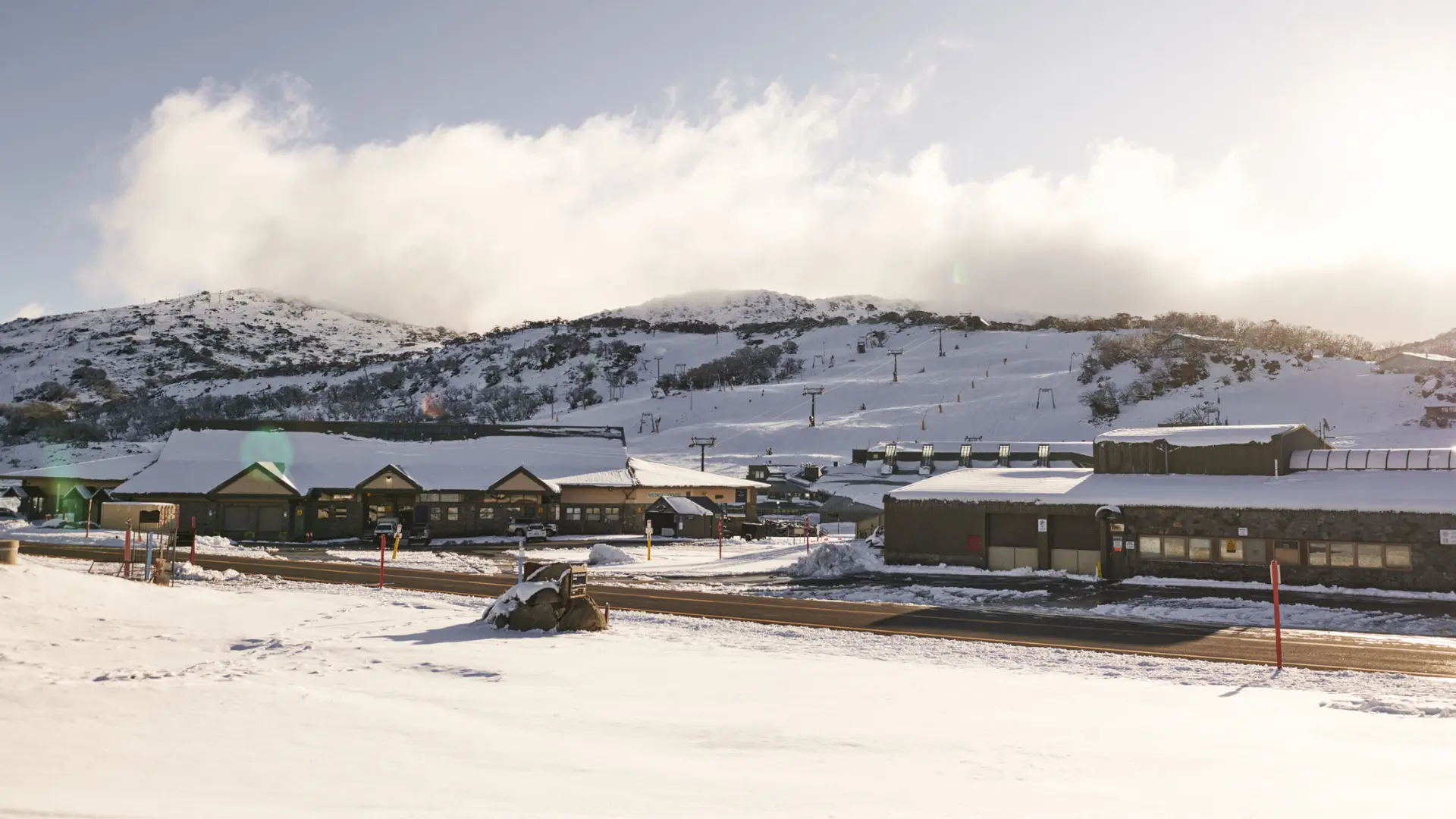 Snow-covered ski area with small lodge buildings, chairlifts on the hillside, and low clouds drifting over the mountain under soft sunlight