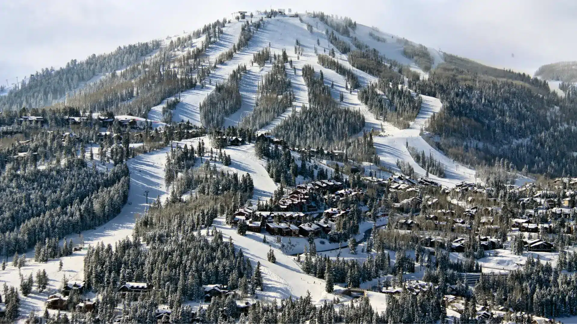Snow-covered ski mountain with groomed slopes, pine forests, and alpine lodges under a bright winter sky