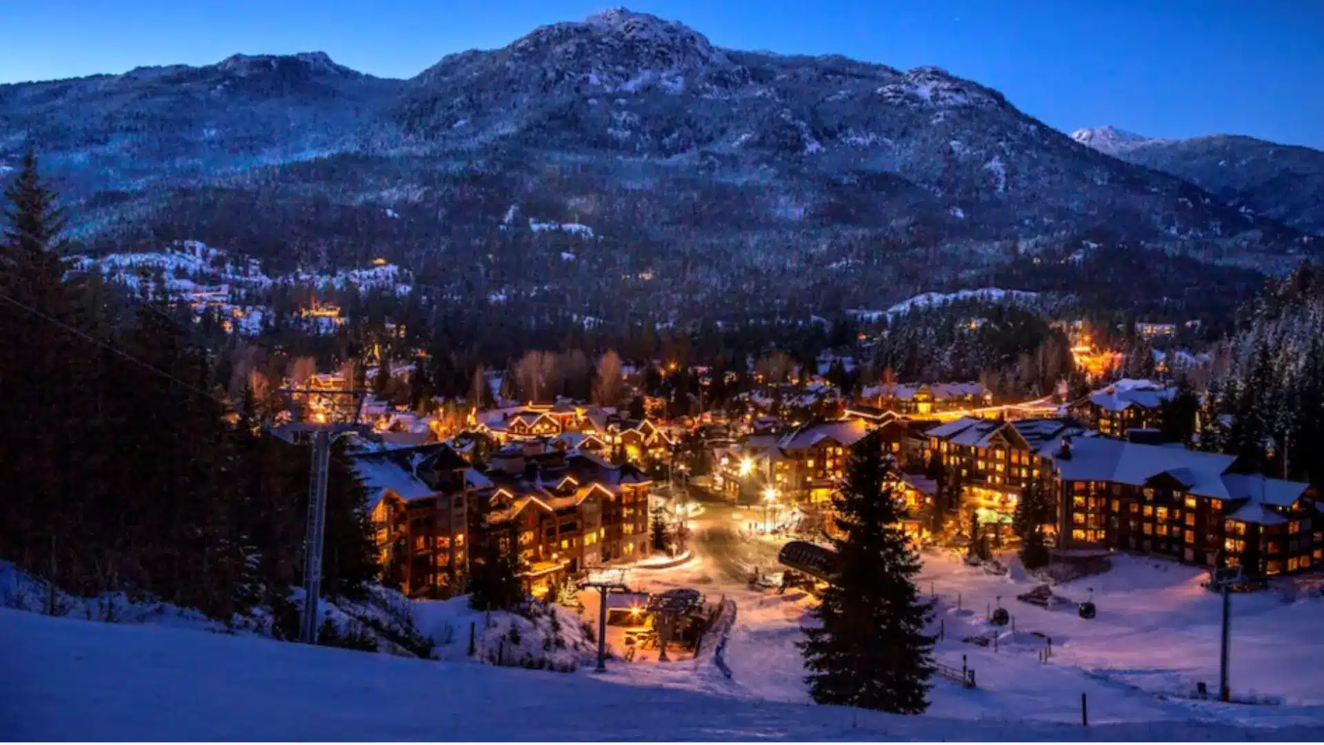 Snow-covered ski resort village at night with warm lights, chairlifts, and forested mountains in the background