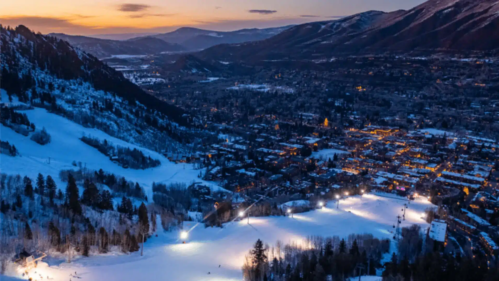Snow-covered ski slopes overlooking a lit mountain town at sunset with surrounding hills and winter landscape