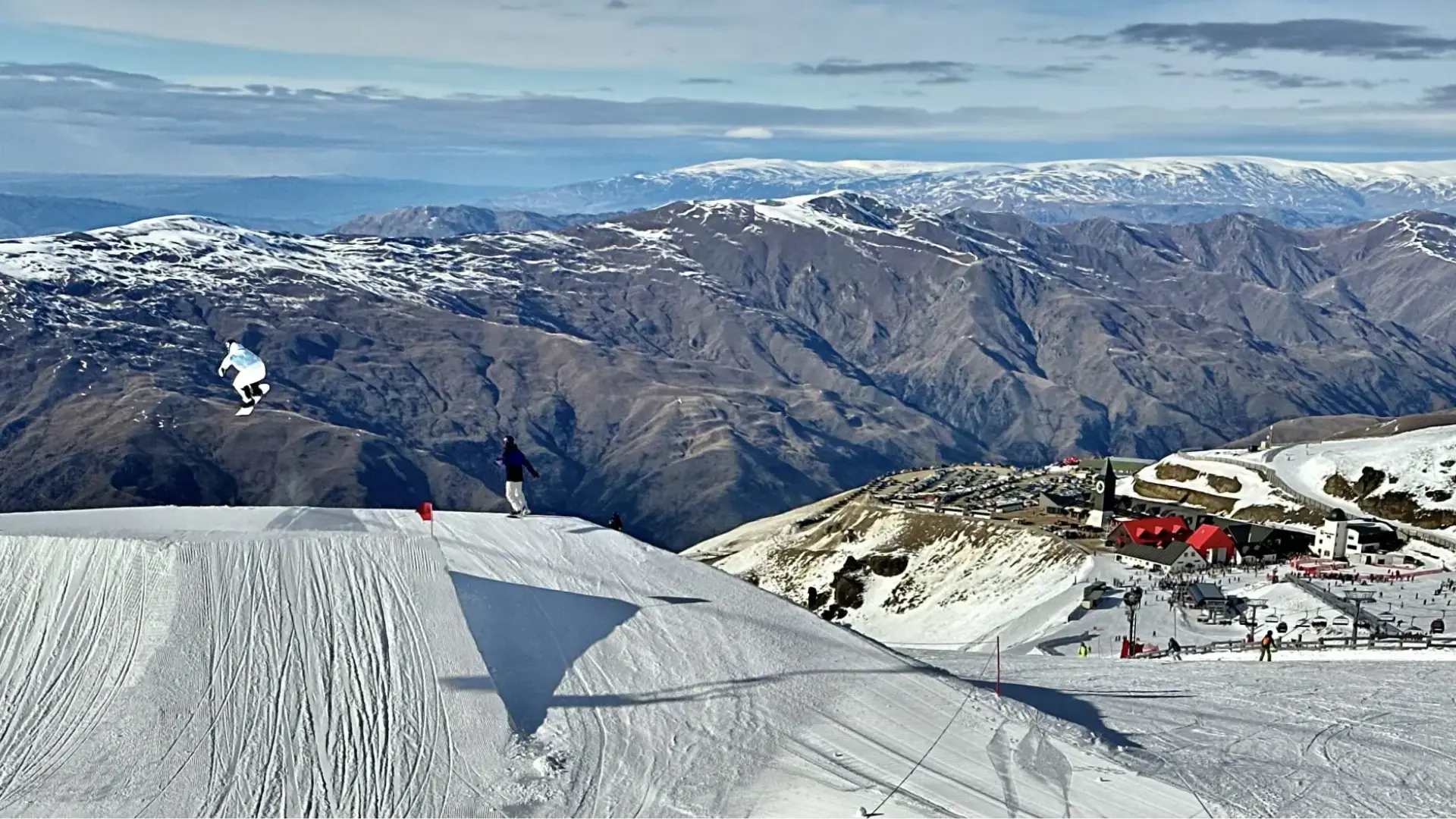 Snowboarder performing a jump on a snowy ridge at a mountain ski resort, with dramatic alpine peaks and buildings in the background