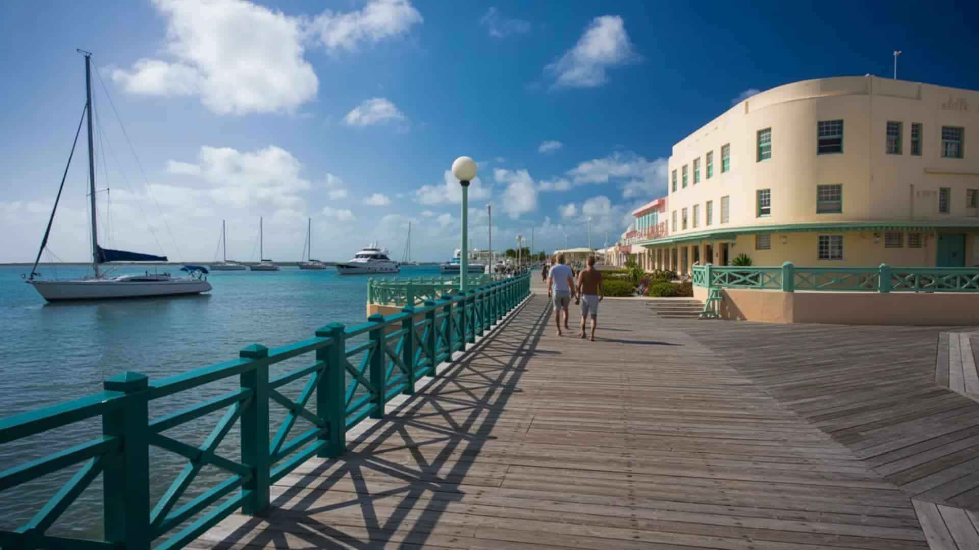Waterfront boardwalk with turquoise railings, boats in the harbor, and people walking beside a light-colored seaside building