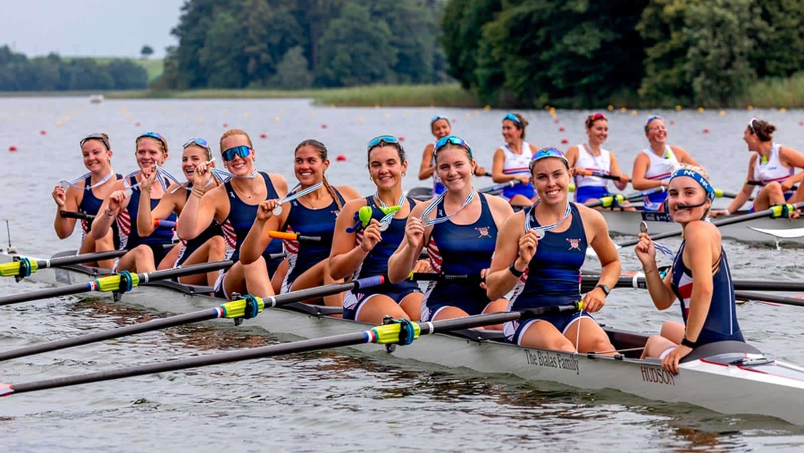 Women’s college rowing team sitting in a racing shell on the water, holding medals after a regatta_