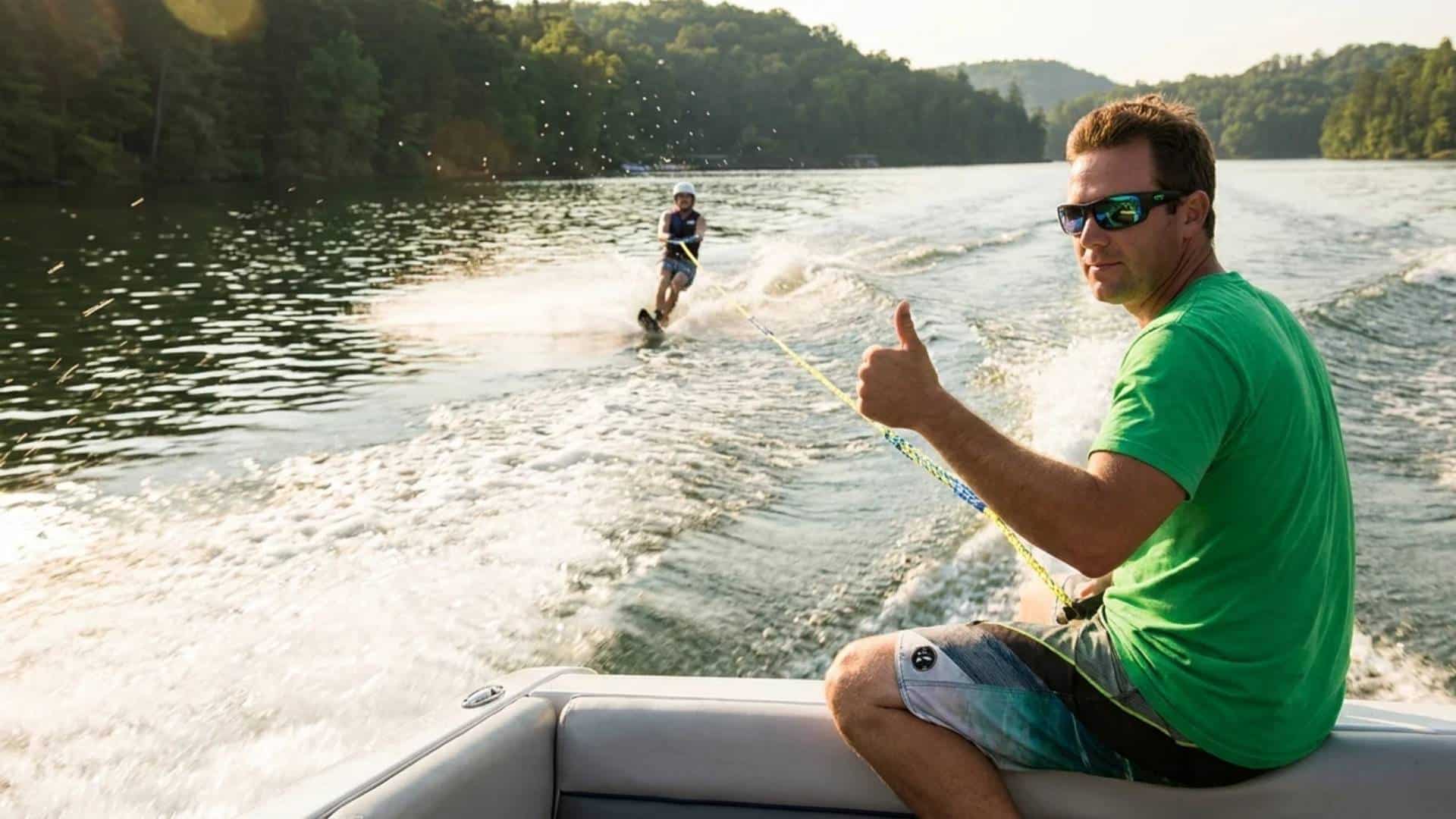 Spotter giving thumbs-up to water skier behind a boat on a calm lake during daytime.