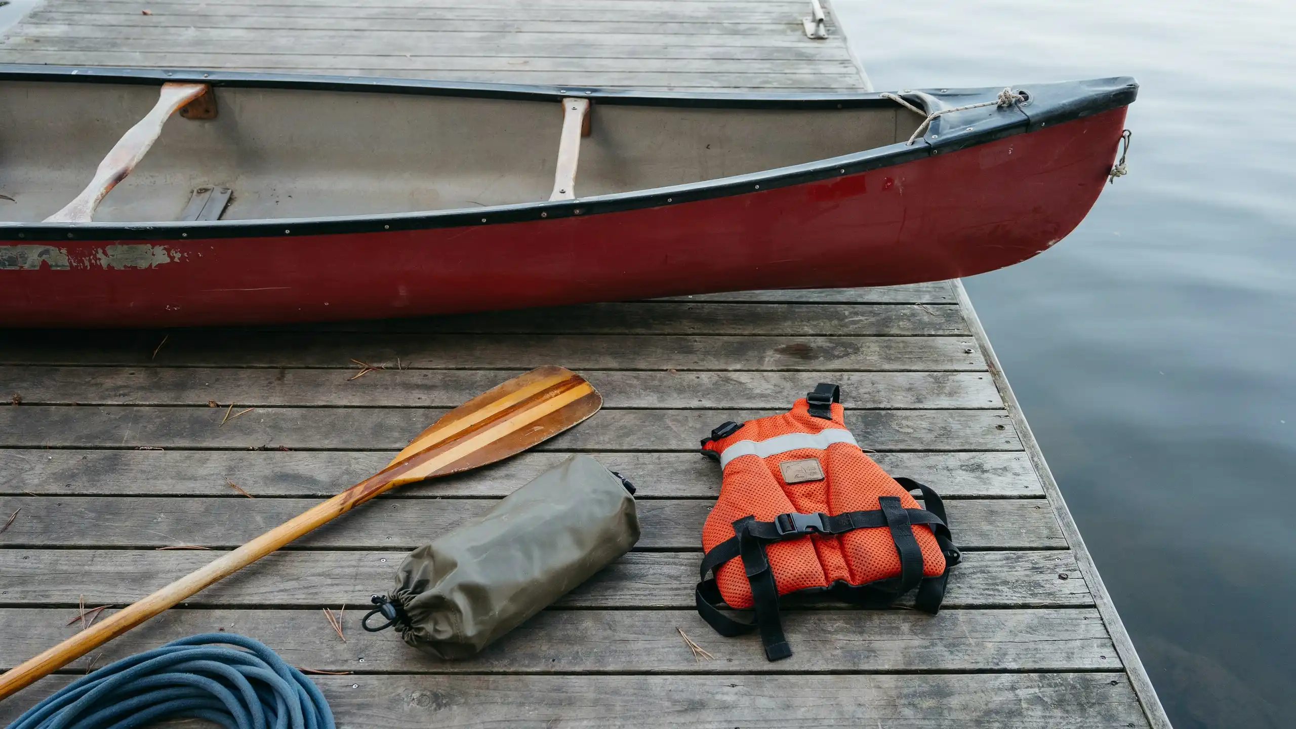 Canoe, wooden paddle, life jacket, dry bag, rope and safety gear laid out on a dock beside calm water ready for a canoe trip.
