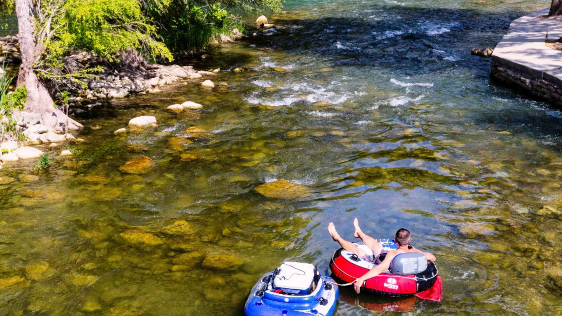 Two people floating in inner tubes on a clear shallow river with rocks visible and gentle current in summer day
