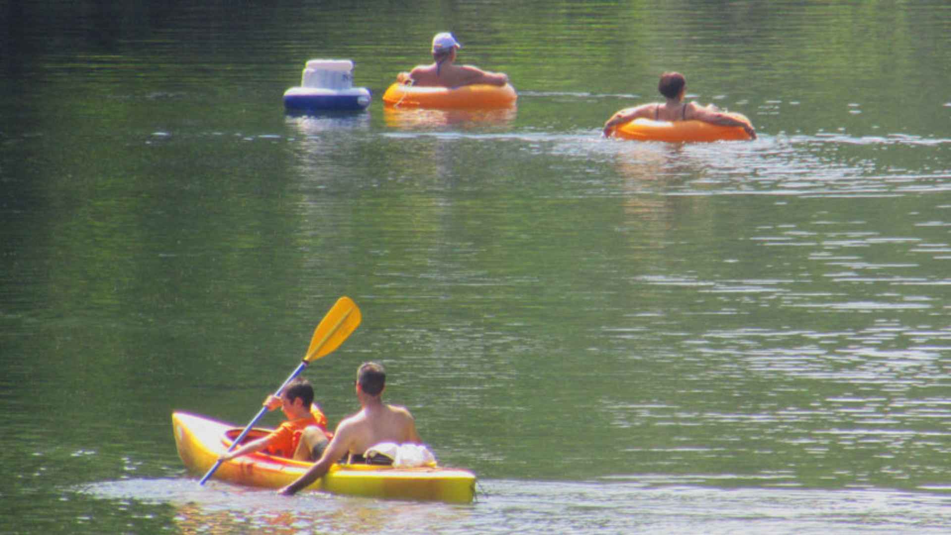 Teo people boating and two people floating in inner tubes on a clear shallow river with rocks visible and gentle current in summer day