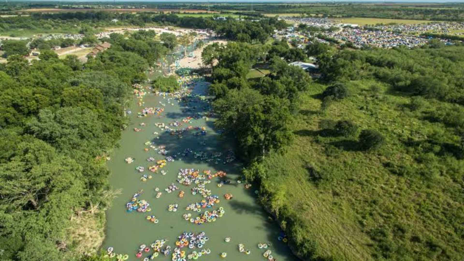 Aerial view of a busy river floating scene with dozens of colorful tubes drifting through green forested landscape and town nearby.