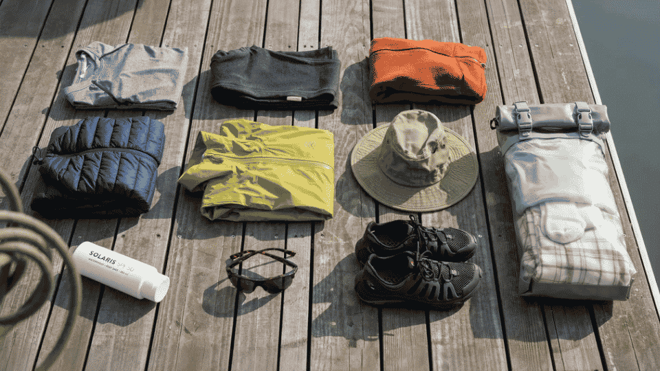 Flat lay of canoe camping clothing and gear including base layers, rain jacket, water shoes, and dry bag on dock.