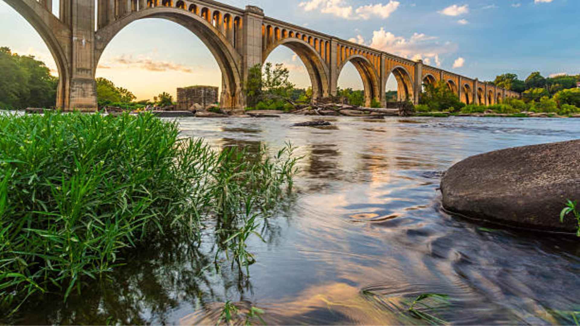 Historic stone arch bridge spanning a wide river at sunset with flowing water, greenery, and rocks along the riverbank.