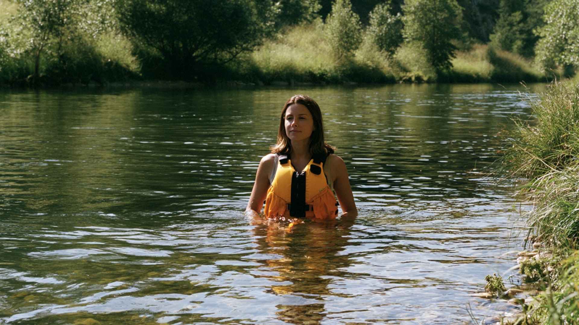 Woman standing in calm inland river water wearing a yellow Level 50 life jacket surrounded by green trees.