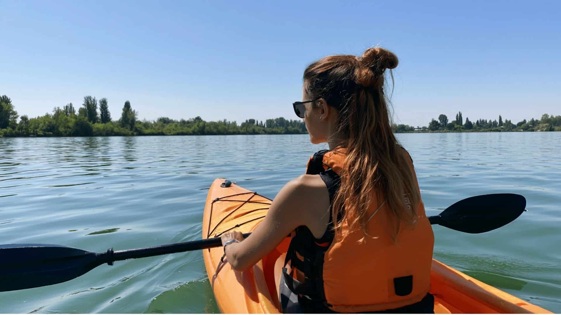 Woman kayaking on calm inland water wearing an orange Level 70 life jacket on a clear sunny day.
